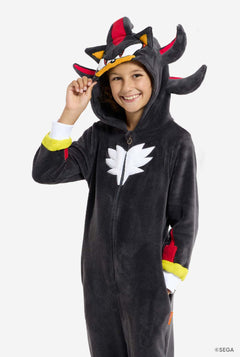 Ein Kind trägt einen schwarzen Plüsch-Onesie für Kinder, der von der beliebten Igel-Figur Shadow inspiriert ist. Er hat rote und schwarze Stacheln an der Kapuze, ein weißes Brustemblem und gelbe Details an den Handgelenken. Der Strampler ist aus weichem Fleecematerial gefertigt und bietet Komfort und ein verspieltes Design für Cosplay, Faulenzen oder thematische Veranstaltungen. Die Person lächelt, während die Kapuze auf ihrem Kopf sitzt. Perfekt für Fans von Spielen oder Zeichentrickfiguren.