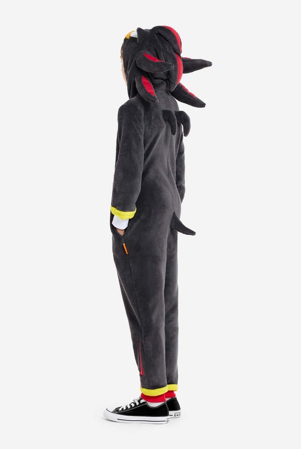 Ein Kind trägt einen plüschigen schwarzen Kinder-Onesie, inspiriert vom beliebten Igel Shadow. Er besticht durch stachelige rote und schwarze Akzente an der Kapuze, ein weißes Brustemblem und gelbe Handgelenksdetails. Der Onesie aus weichem Fleece bietet Komfort und ein verspieltes Design für Cosplay, zum Entspannen oder für Themenveranstaltungen. Seitenansicht. Perfekt für Fans von Videospielen oder Zeichentrickfiguren.