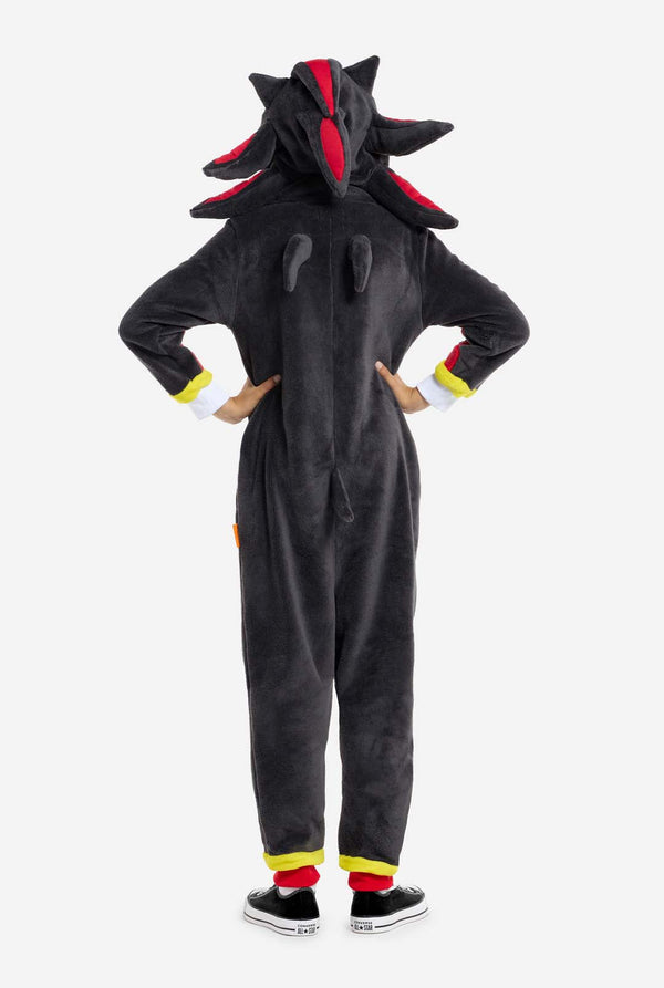 Ein Kind trägt einen plüschigen schwarzen Kinder-Onesie, inspiriert vom beliebten Igel Shadow. Er hat stachelige rote und schwarze Akzente an der Kapuze, ein weißes Brustemblem und gelbe Handgelenksdetails. Der Onesie aus weichem Fleece bietet Komfort und ein verspieltes Design für Cosplay, zum Entspannen oder für Themenveranstaltungen. Rückansicht. Perfekt für Fans von Videospielen oder Zeichentrickfiguren.