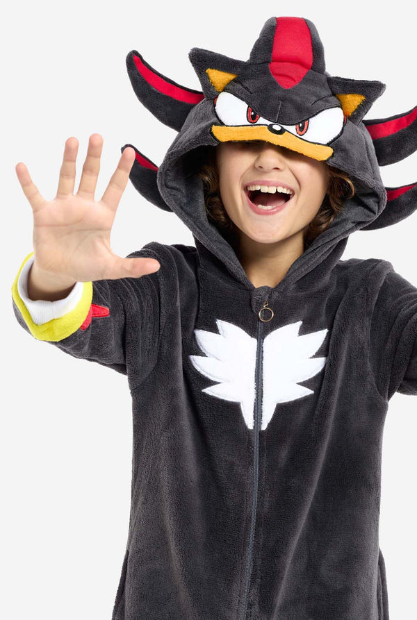Ein Kind trägt einen plüschigen schwarzen Kinder-Onesie, inspiriert vom beliebten Igel Shadow. Er besticht durch stachelige rote und schwarze Akzente an der Kapuze, ein weißes Brustemblem und gelbe Handgelenksdetails. Der Onesie aus weichem Fleece bietet Komfort und ein verspieltes Design für Cosplay, zum Entspannen oder für Themenveranstaltungen. Nahaufnahme mit der Kapuze über den Augen. Perfekt für Fans von Videospielen oder Zeichentrickfiguren.