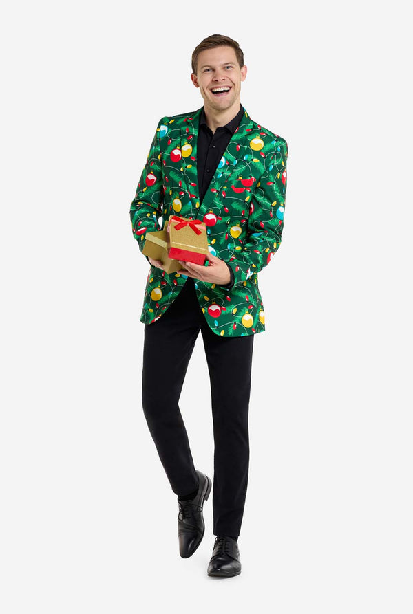Mann trägt einen grünen Weihnachtsblazer für Herren mit verziertem Weihnachtsbaum-Aufdruck. Der Mann trägt außerdem eine schwarze Hose und ein schwarzes Herrenhemd. Hält Geschenke.