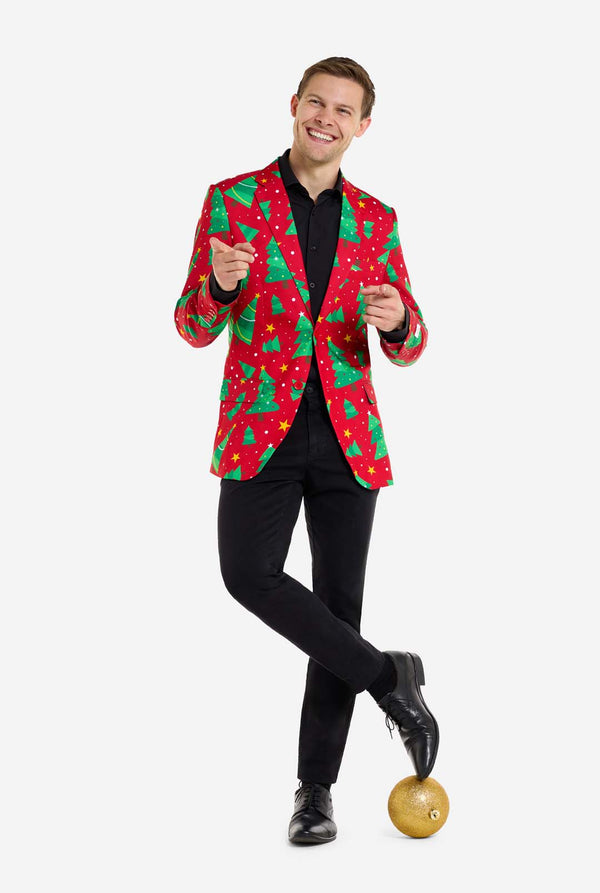 Mann trägt einen roten Weihnachtsblazer mit Weihnachtsbäumen und Sternen. Er trägt außerdem eine schwarze Hose und ein schwarzes Hemd. Er steht mit einem Fuß auf der Weihnachtskugel. Ganzkörperaufnahme.