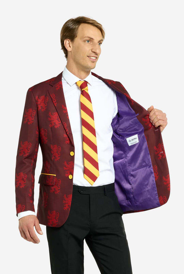 Bild mit hellgrauem Hintergrund und einem Mann, der einen Harry-Potter-Anzug im Gryffindor-Stil trägt. Der Anzug besteht aus einem roten Blazer mit Gryffindor-Logo und hellroten Löwen-Allover-Print, schwarzen Hosen und einer rot-gelb diagonal gestreiften Krawatte. Der Mann hält eine Seite des Blazers geöffnet und zeigt das lila Innenfutter.