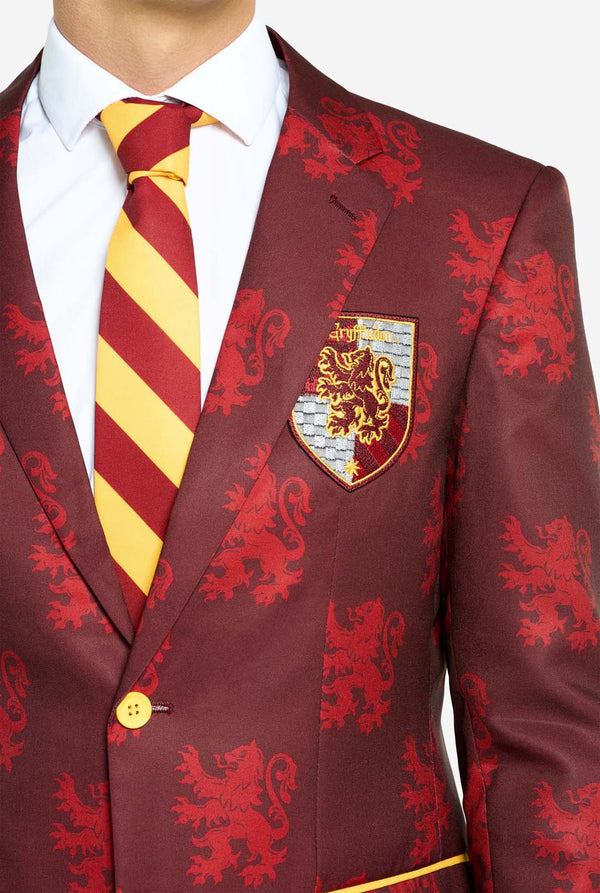 Bild mit hellgrauem Hintergrund und einem Mann, der einen Harry-Potter-Anzug im Gryffindor-Stil trägt. Der Anzug besteht aus einem roten Blazer mit Gryffindor-Logo und einem hellroten Löwen-Allover-Print, schwarzen Hosen und einer rot-gelb diagonal gestreiften Krawatte. Nahaufnahme des Oberkörpers.