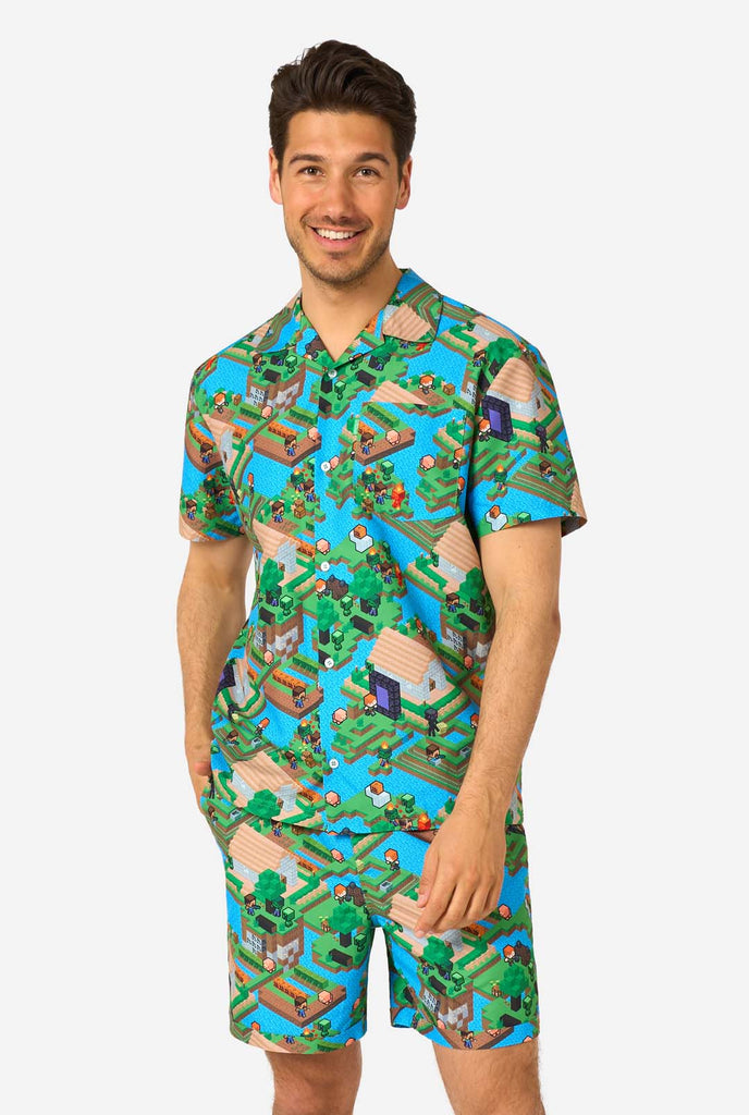 Ein Mann trägt ein buntes Sommeroutfit mit einem Minecraft-Overworld-Motiv, das pixelige Landschaften, Charaktere und Elemente aus dem ikonischen Spiel zeigt. Das kurzärmelige Hemd und die passenden Shorts heben ein lebhaftes Design hervor, das von der Minecraft-Welt inspiriert ist.