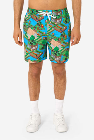 Ein Mann trägt ein buntes Sommeroutfit mit einem Minecraft-Overworld-Motiv, das pixelige Landschaften, Charaktere und Elemente aus dem ikonischen Spiel zeigt. Das kurzärmelige Hemd und die passenden Shorts heben ein lebhaftes Design hervor, das von der Minecraft-Welt inspiriert ist. Nahaufnahme der Shorts.