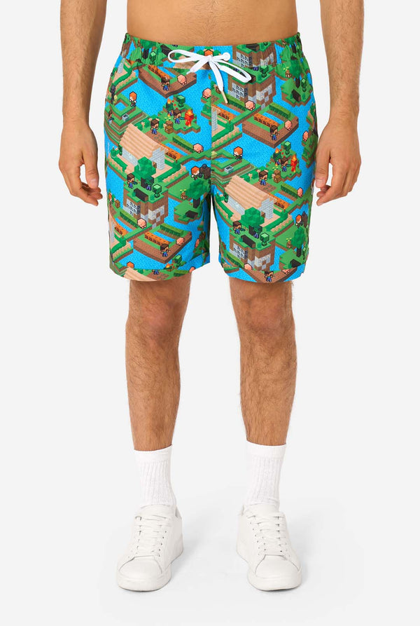 Ein Mann trägt ein buntes Sommeroutfit mit einem Minecraft-Overworld-Motiv, das pixelige Landschaften, Charaktere und Elemente aus dem ikonischen Spiel zeigt. Das kurzärmelige Hemd und die passenden Shorts heben ein lebhaftes Design hervor, das von der Minecraft-Welt inspiriert ist. Nahaufnahme der Shorts.