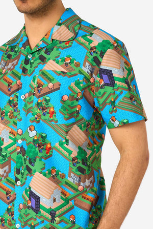 Ein Mann trägt ein buntes Sommeroutfit mit einem Minecraft-Overworld-Motiv, das pixelige Landschaften, Charaktere und Elemente aus dem ikonischen Spiel zeigt. Das kurzärmelige Hemd und die passenden Shorts heben ein lebhaftes Design hervor, das von der Minecraft-Welt inspiriert ist. Nahaufnahme der Brust.