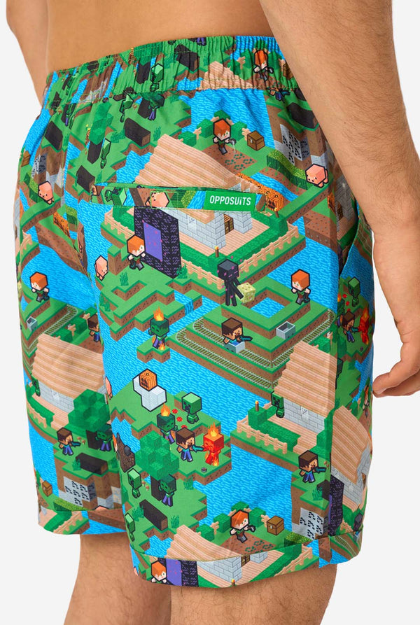 Ein Mann trägt ein buntes Sommeroutfit mit einem Minecraft-Overworld-Motiv, das pixelige Landschaften, Charaktere und Elemente aus dem ikonischen Spiel zeigt. Das kurzärmelige Hemd und die passenden Shorts heben ein lebhaftes Design hervor, das von der Minecraft-Welt inspiriert ist. Nahaufnahme der Shorts von hinten.