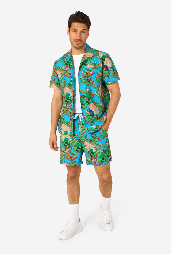 Ein Mann trägt ein buntes Sommeroutfit mit einem Minecraft-Overworld-Motiv, das pixelige Landschaften, Charaktere und Elemente aus dem ikonischen Spiel zeigt. Das kurzärmelige Hemd und die passenden Shorts heben ein lebhaftes Design hervor, das von der Minecraft-Welt inspiriert ist. Ganzkörperbild.