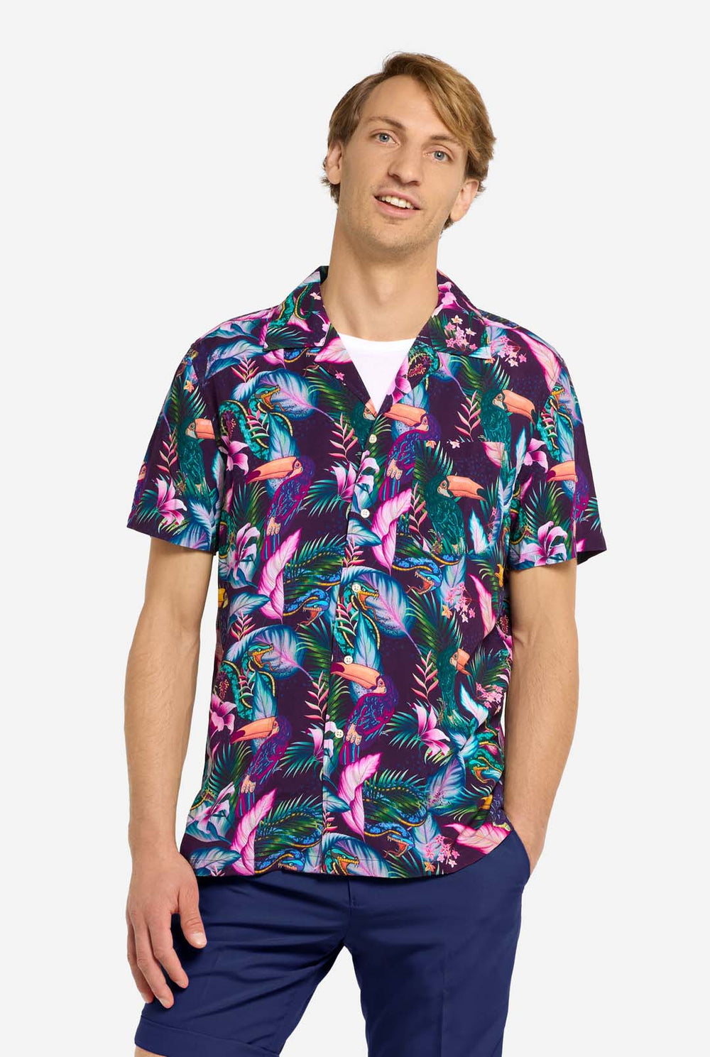 Ein Mann trägt ein lila Hawaiihemd mit tropischem Muster, das an das Nachtleben im Dschungel erinnert. Das Muster besteht aus Papageien, Schlangen, Blättern und Blumen. Dazu trägt er lila Shorts. Er lächelt in die Kamera und hat eine Hand in der Tasche.