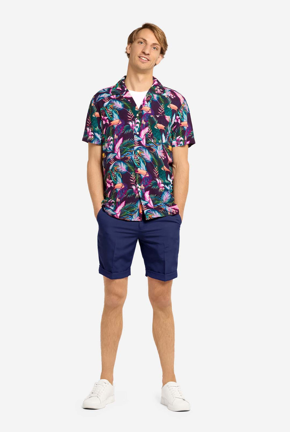 Ein Mann trägt ein lila Hawaiihemd mit tropischem Muster, das an das Nachtleben im Dschungel erinnert. Das Muster besteht aus Papageien, Schlangen, Blättern und Blumen. Dazu trägt er lila Shorts. Er lächelt in die Kamera und hat eine Hand in der Tasche.