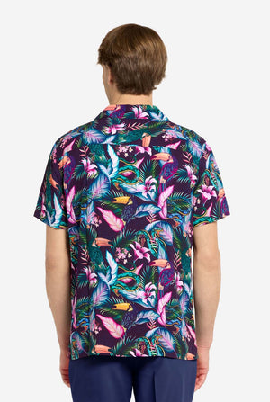 Ein Mann trägt ein lila Hawaiihemd mit tropischem Print, der an das Nachtleben im Dschungel erinnert. Der Print besteht aus Papageien, Schlangen, Blättern und Blumen. Dazu trägt er lila Shorts. Er lächelt in die Kamera und hat eine Hand in der Tasche. Foto von hinten.