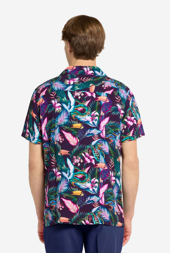 Ein Mann trägt ein lila Hawaiihemd mit tropischem Print, der an das Nachtleben im Dschungel erinnert. Der Print besteht aus Papageien, Schlangen, Blättern und Blumen. Dazu trägt er lila Shorts. Er lächelt in die Kamera und hat eine Hand in der Tasche. Foto von hinten.