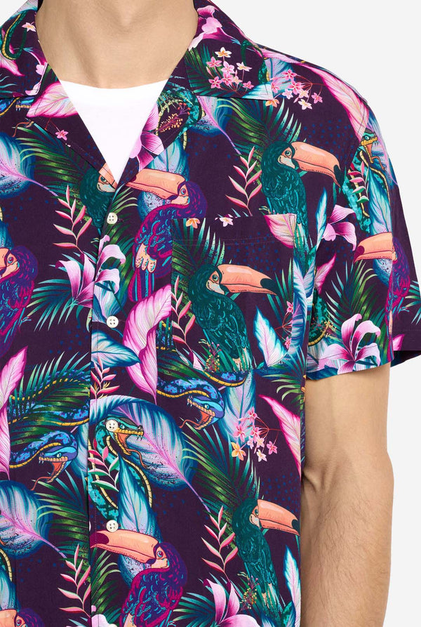 Ein Mann trägt ein lila Hawaiihemd mit tropischem Print, der an Jungle Night Live erinnert. Der Print besteht aus Papageien, Schlangen, Blättern und Blumen. Dazu trägt er rosa Shorts. Er lächelt in die Kamera und hält eine Hand in der Tasche. Nahaufnahme.
