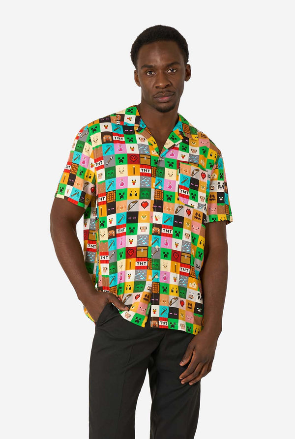 Mann, der ein farbenfrohes Minecraft-Hawaiihemd mit pixeligen Blöcken, Charakteren und Gegenständen aus dem Spiel trägt, gepaart mit einer schwarzen Hose, steht vor einem schlichten weißen Hintergrund.