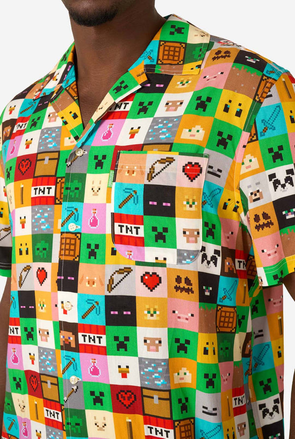 Mann, der ein farbenfrohes Minecraft-Hawaiihemd mit pixeligen Blöcken, Charakteren und Gegenständen aus dem Spiel trägt, gepaart mit einer schwarzen Hose, steht vor einem schlichten weißen Hintergrund. Nahaufnahme der Brust