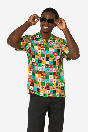 Mann, der ein farbenfrohes Minecraft-Hawaiihemd mit pixeligen Blöcken, Charakteren und Gegenständen aus dem Spiel trägt, gepaart mit einer schwarzen Hose, steht vor einem schlichten weißen Hintergrund. Sonnenbrille tragen