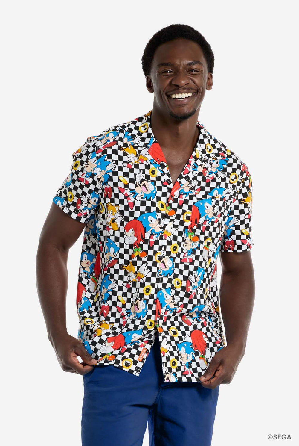 Ein Mann trägt ein schwarz-weiß kariertes Hawaiihemd mit Figuren aus der Sonic the Hedgehog-Reihe und goldenen Ringen darauf. Er lächelt in die Kamera.