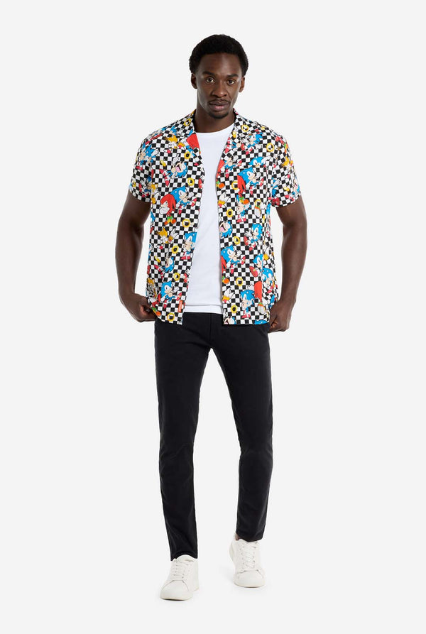 Ein Mann trägt ein schwarz-weiß kariertes Hawaiihemd mit Figuren aus der Sonic the Hedgehog-Reihe und goldenen Ringen. Dazu trägt er eine schwarze Hose. Er blickt in die Kamera und hält den unteren Teil des Hemdes fest.