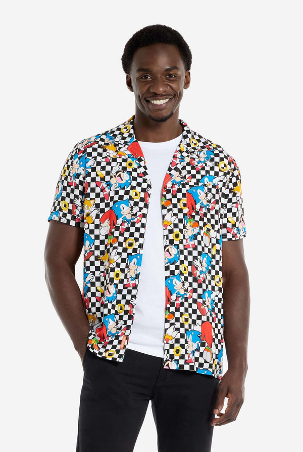 Ein Mann trägt ein schwarz-weiß kariertes Hawaiihemd mit Figuren aus der Sonic the Hedgehog-Reihe und goldenen Ringen. Dazu trägt er eine schwarze Hose. Er lächelt in die Kamera und hält den unteren Teil des Hemdes.
