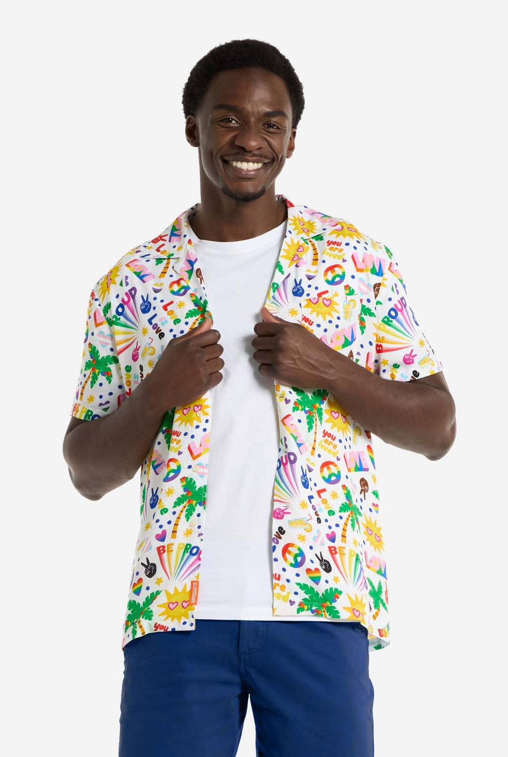 Ein Mann trägt ein weißes Hawaii-Hemd mit einem Regenbogen-Pride-Aufdruck. Der Aufdruck besteht aus Friedenssymbolen, Herzen und anderen Symbolen. Er trägt außerdem eine blaue Hose. Er lächelt in die Kamera und hält die Seiten des Hemdes fest.