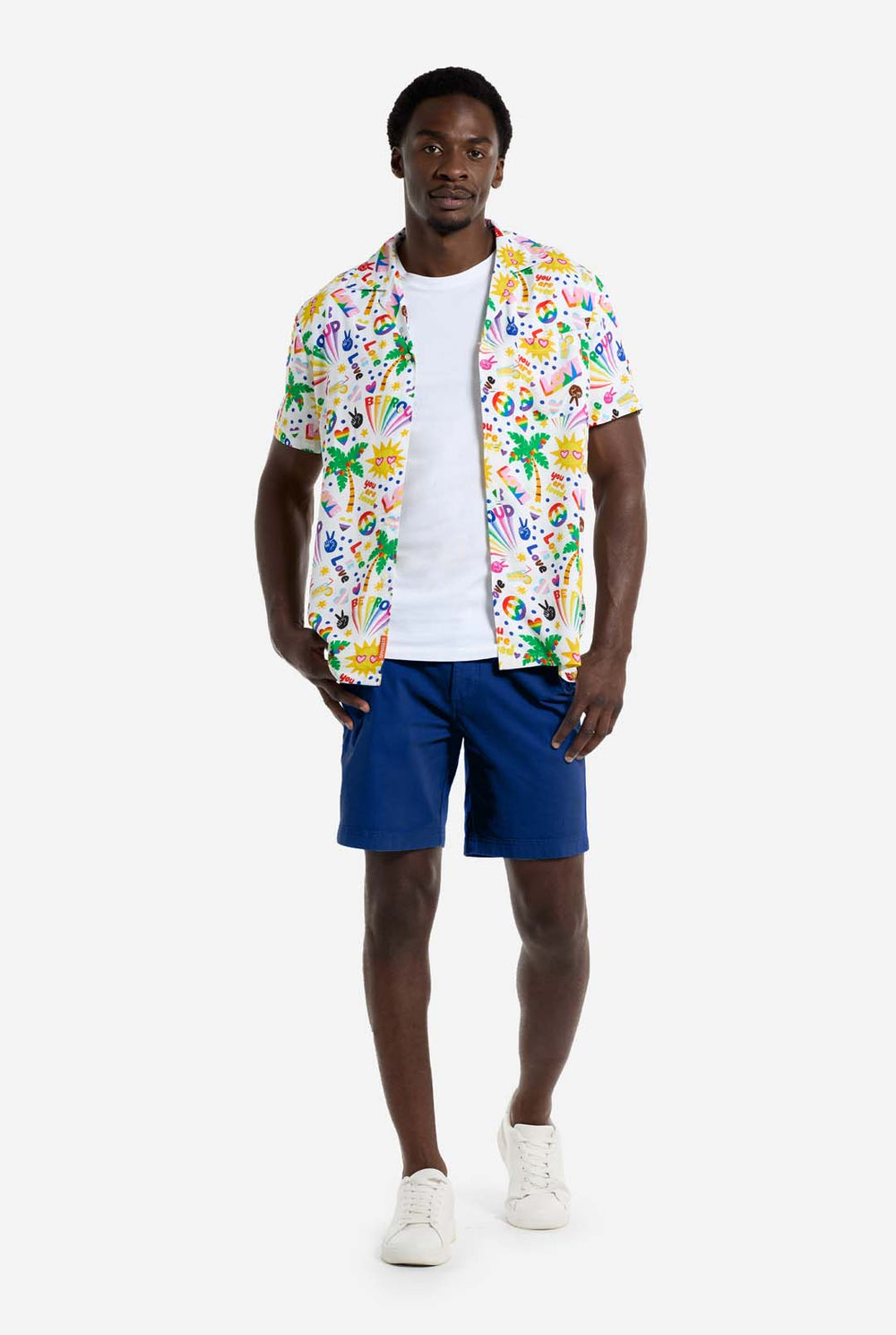 Ein Mann trägt ein weißes Hawaii-Hemd mit einem Regenbogen-Pride-Aufdruck. Der Aufdruck besteht aus Friedenssymbolen, Herzen und anderen Symbolen. Er trägt außerdem eine blaue Hose. Er lächelt in die Kamera und hält die Seiten des Hemdes fest.