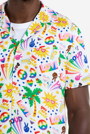 Ein Mann trägt ein weißes Hawaii-Hemd mit einem Regenbogen-Pride-Aufdruck. Der Aufdruck besteht aus Friedenssymbolen, Herzen und anderen Symbolen. Nahaufnahme der Brust.
