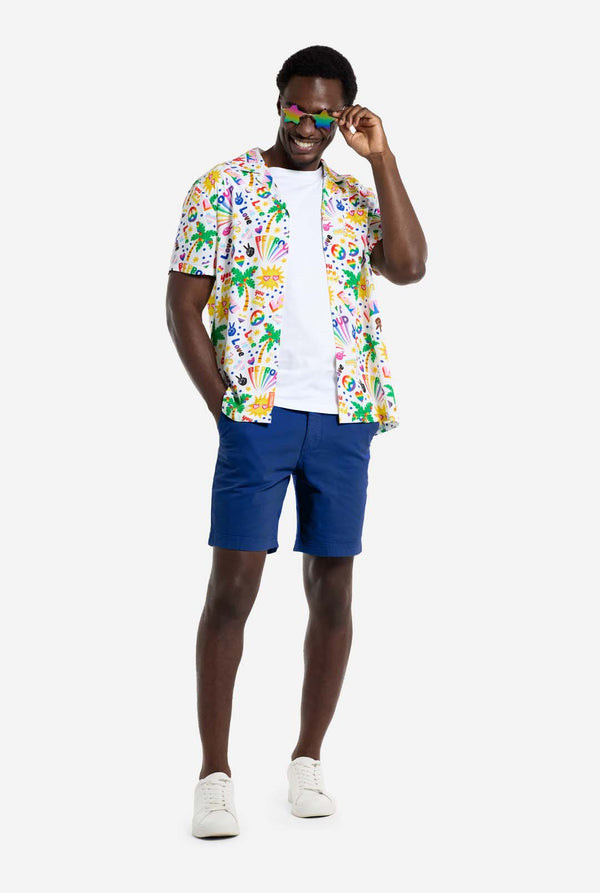 Ein Mann trägt ein weißes Hawaii-Hemd mit einem Regenbogen-Pride-Aufdruck. Der Aufdruck besteht aus Friedenssymbolen, Herzen und anderen Symbolen. Er trägt außerdem eine blaue Hose. Er lächelt und trägt eine sternförmige Sonnenbrille.
