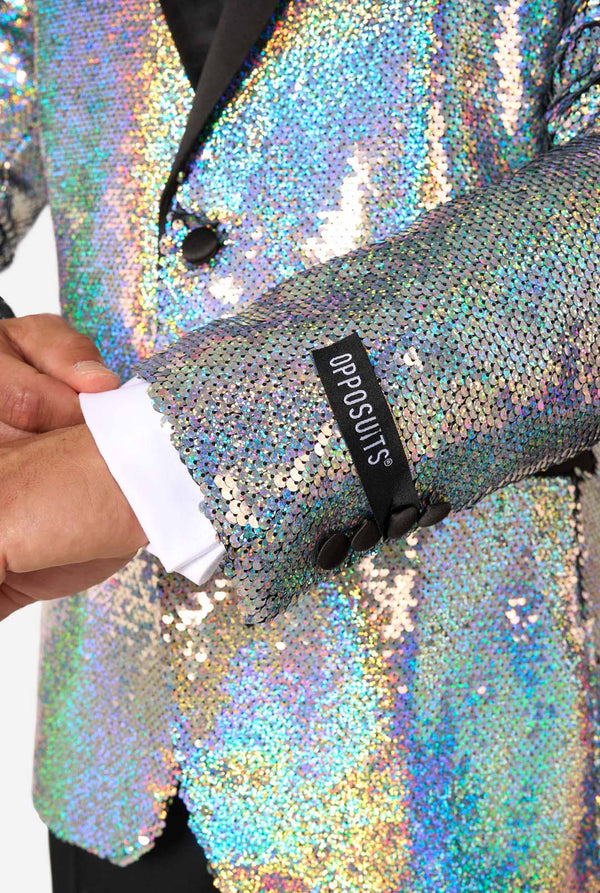 Mann, der einen holographischen silbernen Paillettentuxedo mit schwarzen Satin -Repfels trägt, gepaart mit einem weißen Hemd, einer schwarzen Fliege und einer schwarzen Hose.
