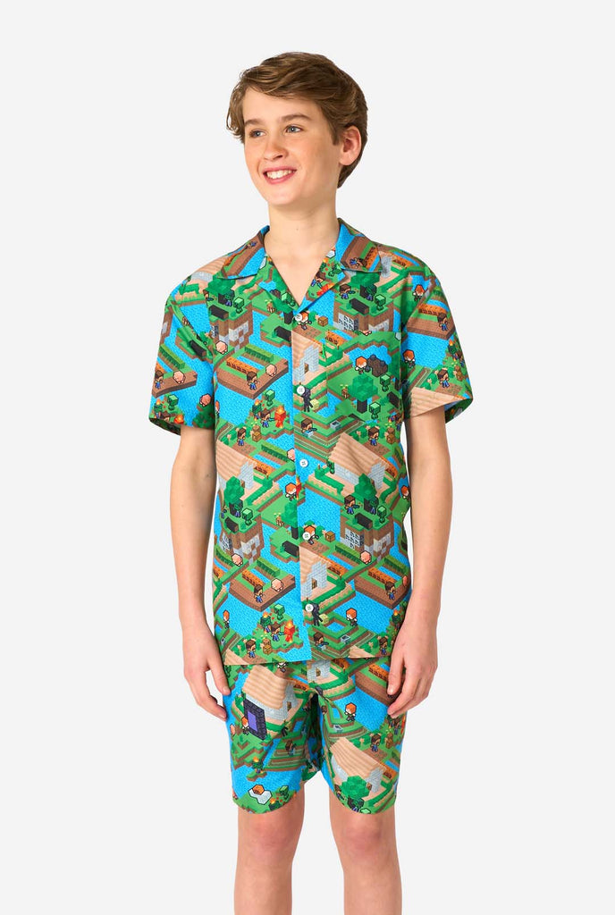 Ein Teenager-Junge trägt ein buntes Sommeroutfit mit einem Minecraft-Overworld-Motiv, das pixelige Landschaften, Charaktere und Elemente aus dem ikonischen Spiel zeigt. Das kurzärmelige Hemd und die passenden Shorts heben ein lebhaftes Design hervor, das von der Minecraft-Welt inspiriert ist.