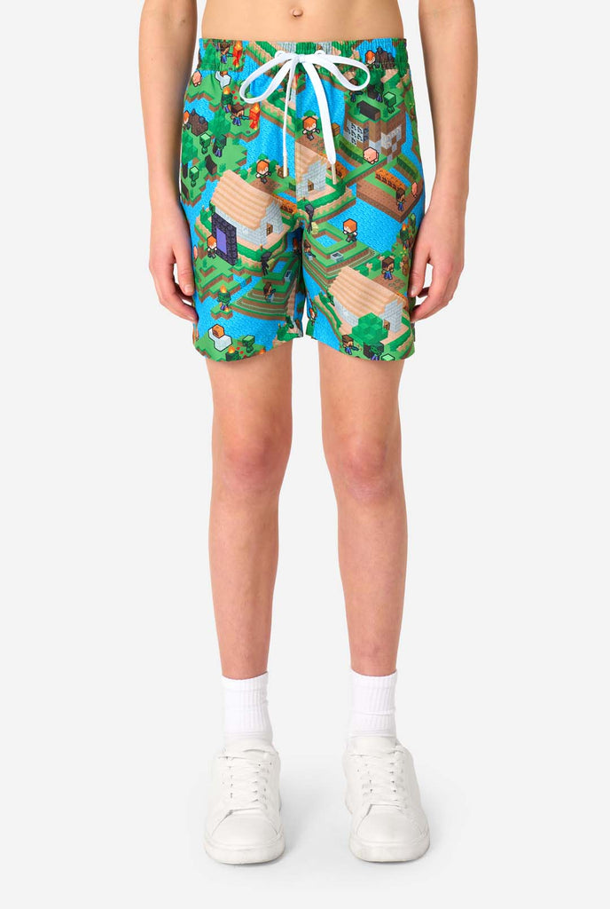 Ein Teenager-Junge trägt ein buntes Sommeroutfit mit einem Minecraft-Overworld-Motiv, das pixelige Landschaften, Charaktere und Elemente aus dem ikonischen Spiel zeigt. Das kurzärmelige Hemd und die passenden Shorts heben ein lebhaftes Design hervor, das von der Minecraft-Welt inspiriert ist. Nahaufnahme der Shorts.