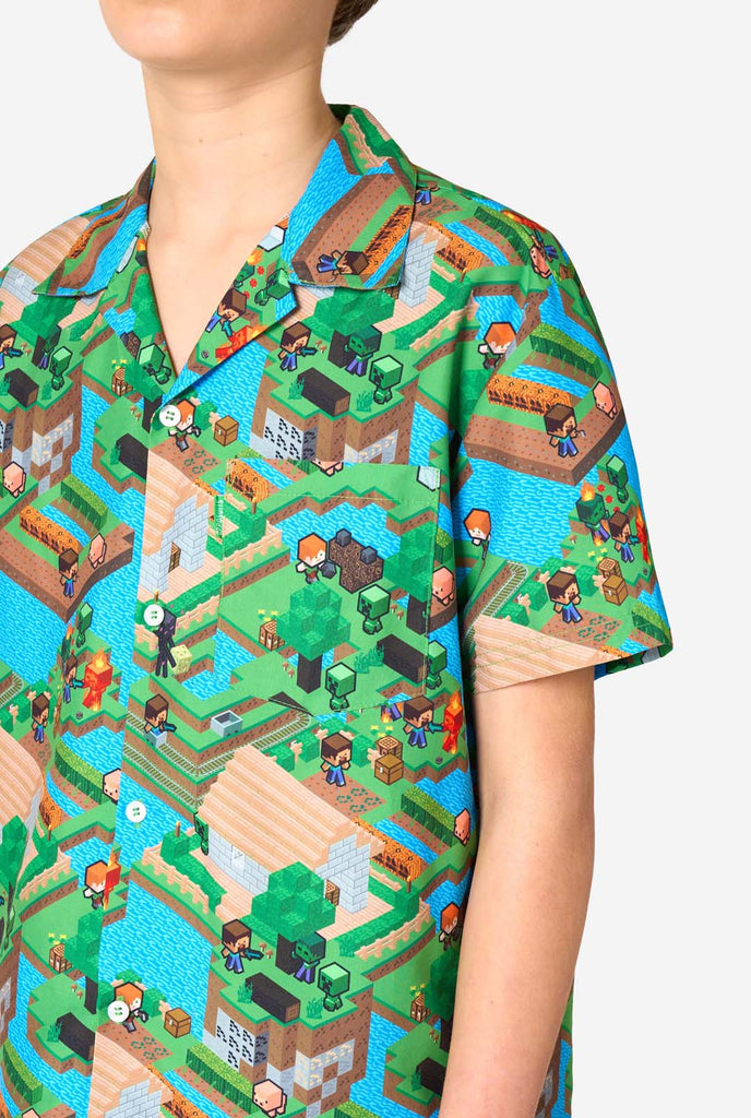 Ein Teenager-Junge trägt ein buntes Sommeroutfit mit einem Minecraft-Overworld-Motiv, das pixelige Landschaften, Charaktere und Elemente aus dem ikonischen Spiel zeigt. Das kurzärmelige Hemd und die passenden Shorts heben ein lebhaftes Design hervor, das von der Minecraft-Welt inspiriert ist. Fokus auf die Brust.