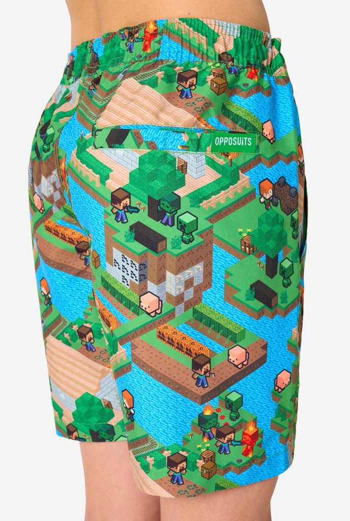 Ein Teenager-Junge trägt ein buntes Sommeroutfit mit einem Minecraft-Overworld-Motiv, das pixelige Landschaften, Charaktere und Elemente aus dem ikonischen Spiel zeigt. Das kurzärmelige Hemd und die passenden Shorts heben ein lebhaftes Design hervor, das von der Minecraft-Welt inspiriert ist. Fokus auf die Shorts von hinten.