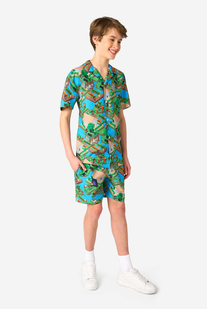 Ein Teenager-Junge trägt ein buntes Sommeroutfit mit einem Minecraft-Overworld-Motiv, das pixelige Landschaften, Charaktere und Elemente aus dem ikonischen Spiel zeigt. Das kurzärmelige Hemd und die passenden Shorts heben ein lebhaftes Design hervor, das von der Minecraft-Welt inspiriert ist. Ganzkörperbild.