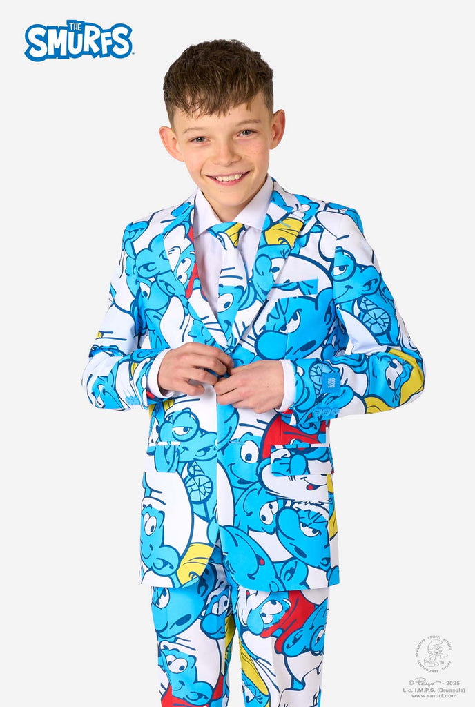 Teenager-Junge trägt einen lebhaften Schlumpf-Kinderanzug mit bunten Cartoon-Motiven in Blau, Rot, Gelb und Weiß, kombiniert mit passender Krawatte und weißem Hemd, lächelt vor einem schlichten Hintergrund.