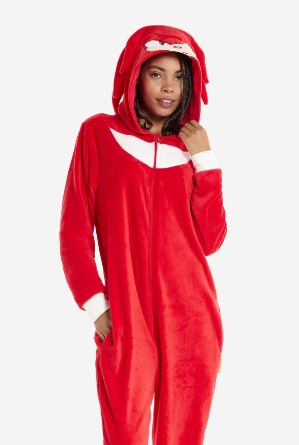 Eine Frau trägt einen leuchtend roten Knuckles the Echidna-Einteiler für Erwachsene, ein kuscheliges und verspieltes Outfit, inspiriert von der berühmten Figur aus der Sonic the Hedgehog-Reihe. Der Einteiler zeigt Knuckles' charakteristischen weißen Brustaufnäher und eine Kapuze, die Knuckles' Gesicht mit seinen ausdrucksstarken Augen und langen roten Dreadlocks zeigt. Der Reißverschluss vorne ermöglicht bequemes Anziehen. 