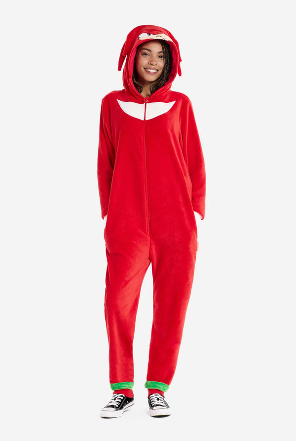 Eine Frau trägt einen leuchtend roten Knuckles the Echidna-Onesie für Erwachsene, ein kuscheliges und verspieltes Outfit, inspiriert von der berühmten Figur aus der Sonic the Hedgehog-Reihe. Der Onesie zeigt Knuckles' charakteristischen weißen Brustaufnäher und eine Kapuze, die Knuckles' Gesicht mit seinen ausdrucksstarken Augen und langen roten Dreadlocks zur Schau stellt. Der Reißverschluss vorne ermöglicht bequemes Anziehen, und die Frau steht mit den Händen in den Seitentaschen. 