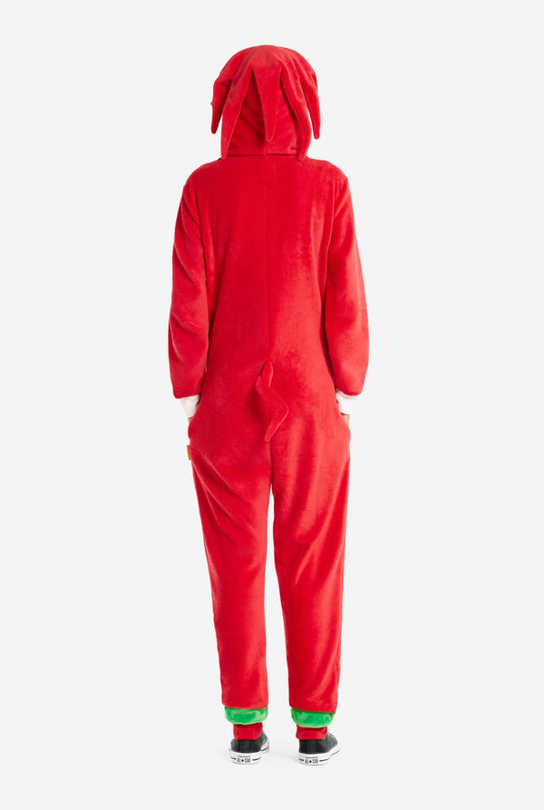 Eine Frau trägt einen leuchtend roten Knuckles the Echidna-Onesie für Erwachsene, ein kuscheliges und verspieltes Outfit, inspiriert von der berühmten Figur aus der Sonic the Hedgehog-Reihe. Der Onesie zeigt Knuckles' charakteristischen weißen Brustaufnäher und eine Kapuze, die Knuckles' Gesicht mit seinen ausdrucksstarken Augen und langen roten Dreadlocks zur Schau stellt. Die Frau steht mit den Händen in den Seitentaschen. 