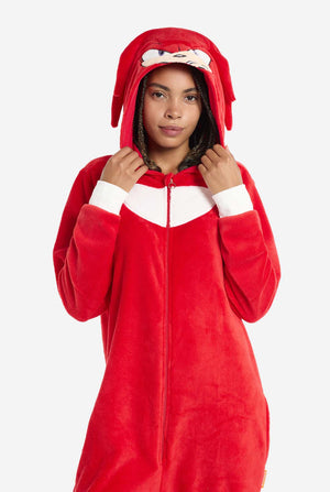 Eine Frau trägt einen leuchtend roten Knuckles the Echidna-Onesie für Erwachsene, ein kuscheliges und verspieltes Outfit, inspiriert von der berühmten Figur aus der Sonic the Hedgehog-Reihe. Der Onesie zeigt Knuckles' charakteristischen weißen Brustaufnäher und eine Kapuze, die Knuckles' Gesicht mit seinen ausdrucksstarken Augen und langen roten Dreadlocks zeigt. Der Reißverschluss vorne ermöglicht bequemes Anziehen, und die Frau hält die Kapuze. 