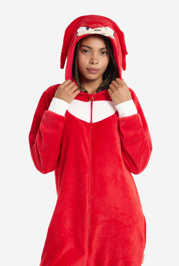 Eine Frau trägt einen leuchtend roten Knuckles the Echidna-Onesie für Erwachsene, ein kuscheliges und verspieltes Outfit, inspiriert von der berühmten Figur aus der Sonic the Hedgehog-Reihe. Der Onesie zeigt Knuckles' charakteristischen weißen Brustaufnäher und eine Kapuze, die Knuckles' Gesicht mit seinen ausdrucksstarken Augen und langen roten Dreadlocks zeigt. Der Reißverschluss vorne ermöglicht bequemes Anziehen, und die Frau hält die Kapuze. 