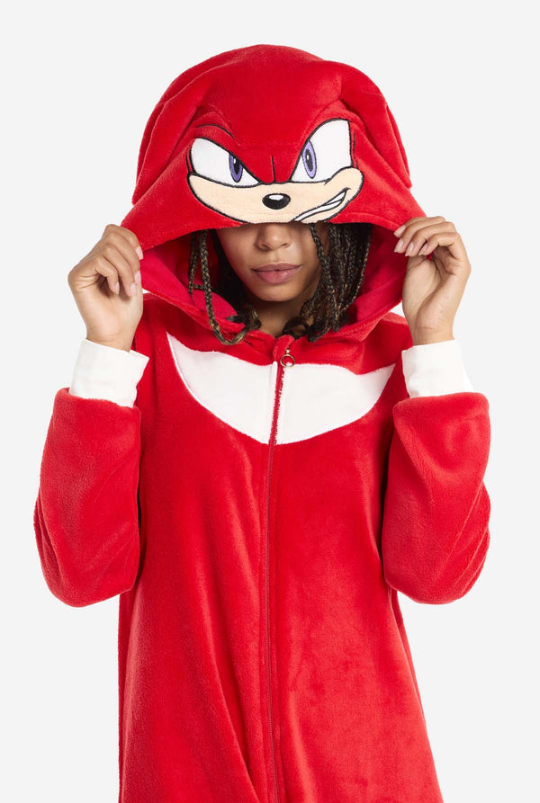 Eine Frau trägt einen leuchtend roten Knuckles the Echidna-Onesie für Erwachsene, ein gemütliches und verspieltes Outfit, inspiriert von der berühmten Figur aus der Sonic the Hedgehog-Serie. Der Onesie zeigt Knuckles' charakteristischen weißen Brustaufnäher und hat eine Kapuze, die Knuckles' Gesicht mit seinen ausdrucksstarken Augen und langen roten Dreadlocks zur Schau stellt. Der Reißverschluss vorne sorgt für einen bequemen Sitz, und die Frau hält die Kapuze über ihre Augen. 