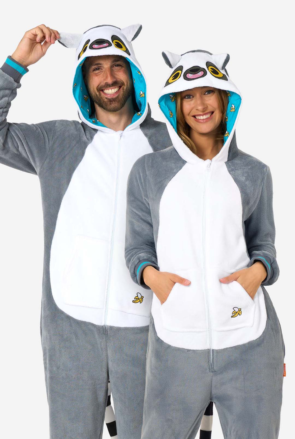 Ein Mann und eine Frau tragen einen grau-weißen Lemur-Onesie für Erwachsene mit verspielten Kapuzendetails und kuschelig weichem Material, perfekt zum Faulenzen oder für Kostümveranstaltungen. Mit schwarzem und weißem Schwanz. Die Kapuze ist auf dem Kopf.