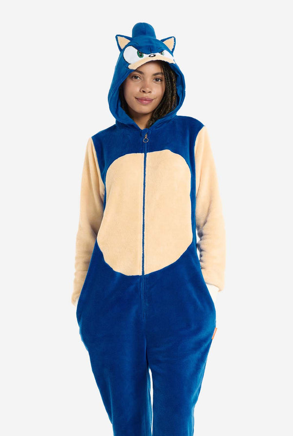 Eine Frau trägt einen kuscheligen, blauen Sonic the Hedgehog-Onesie für Erwachsene. Weiche beigefarbene Flicken auf der Vorderseite und an den Ärmeln imitieren Sonics ikonischen Look. Die Kapuze ist mit Sonics erkennbarem Gesicht gestaltet, einschließlich großer grüner Augen, blauer Stacheln und spitzer Ohren. Der Reißverschluss vorne sorgt für einen bequemen Sitz. Die Frau steht mit den Händen in den Seitentaschen des Onesies. Ihr Gesichtsausdruck ist ruhig und entspannt. 