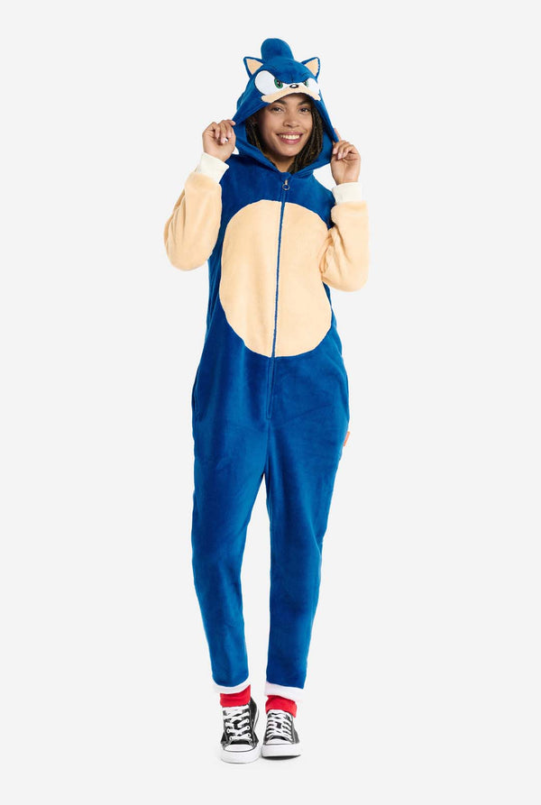 Eine Frau trägt einen kuscheligen, blauen Sonic the Hedgehog-Einteiler für Erwachsene. Weiche beigefarbene Flicken auf der Vorderseite und an den Ärmeln imitieren Sonics ikonischen Look. Die Kapuze des Einteilers ist mit Sonics erkennbarem Gesicht gestaltet, einschließlich großer grüner Augen, blauer Stacheln und spitzer Ohren. Der Reißverschluss vorne sorgt für einen bequemen Sitz, und die Frau hält die Kapuze. Ihr Gesichtsausdruck ist ruhig und entspannt. 