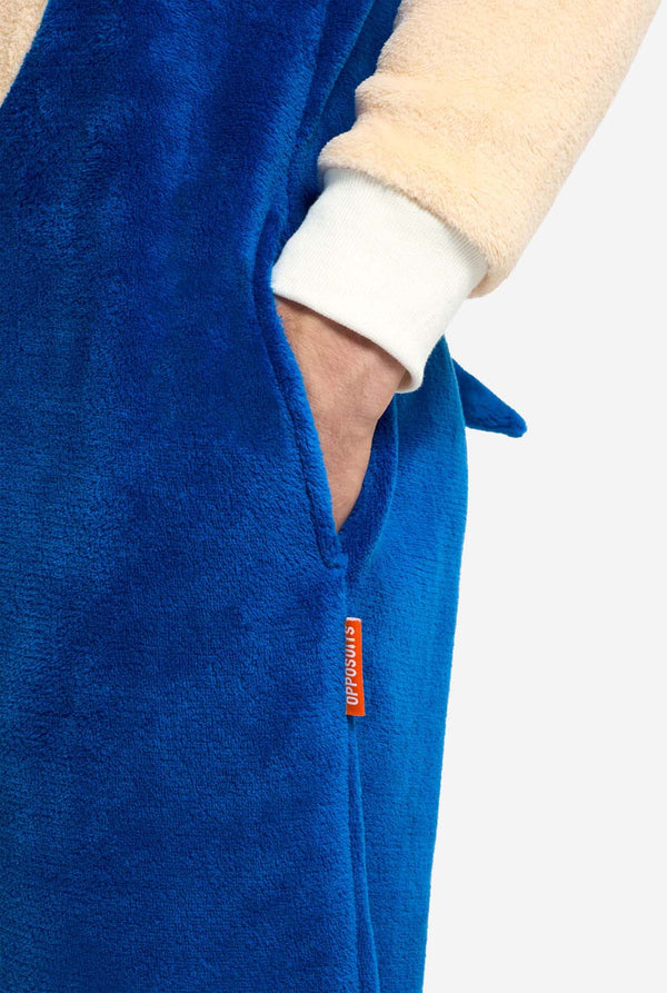 Eine Person trägt einen kuscheligen, blauen Sonic the Hedgehog-Onesie für Erwachsene mit weichen, beigen Ärmeln, die Sonics ikonischen Look nachahmen. Dieser Sonic the Hedgehog-Onesie ist ideal für Cosplay, zum Entspannen oder für Themenveranstaltungen. Nahaufnahme von der Seitentasche mit der Hand in der Tasche.
