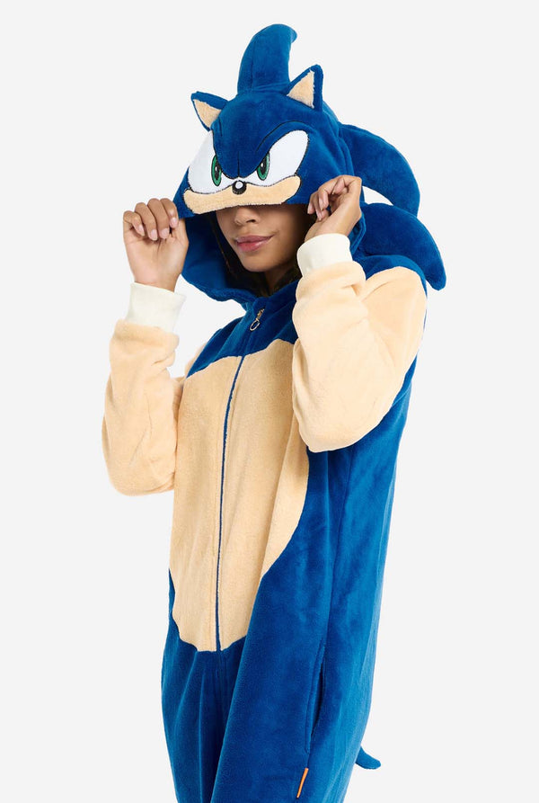 Eine Frau trägt einen kuscheligen, blauen Sonic the Hedgehog-Einteiler für Erwachsene. Weiche beigefarbene Flicken auf der Vorderseite und an den Ärmeln imitieren Sonics ikonischen Look. Die Kapuze des Einteilers ist mit Sonics erkennbarem Gesicht gestaltet, einschließlich großer grüner Augen, blauer Stacheln und spitzer Ohren. Der Reißverschluss vorne sorgt für einen bequemen Sitz, und die Frau hält die Kapuze über die Augen. Ihr Gesichtsausdruck ist ruhig und entspannt. 