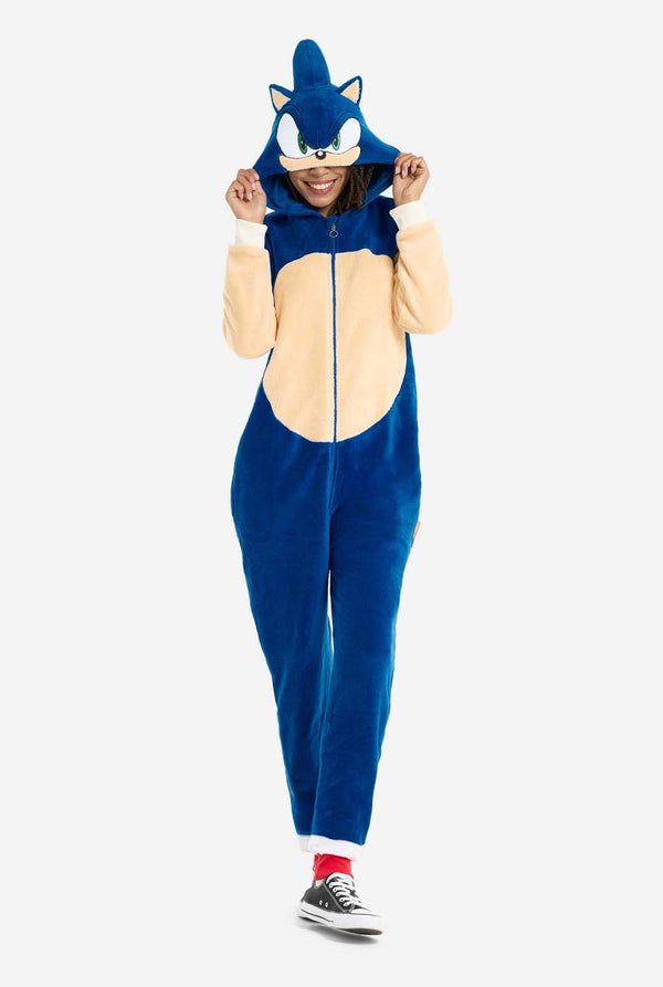 Eine Frau trägt einen kuscheligen, blauen Sonic the Hedgehog-Onesie für Erwachsene. Weiche beigefarbene Flicken auf der Vorderseite und an den Ärmeln imitieren Sonics ikonischen Look. Die Kapuze des Onesie ist mit Sonics bekanntem Gesicht gestaltet, einschließlich großer grüner Augen, blauer Stacheln und spitzer Ohren. Der Reißverschluss vorne sorgt für einen bequemen Sitz, und die Frau hält die Kapuze über die Augen. Ihr Gesichtsausdruck ist ruhig und entspannt. 
