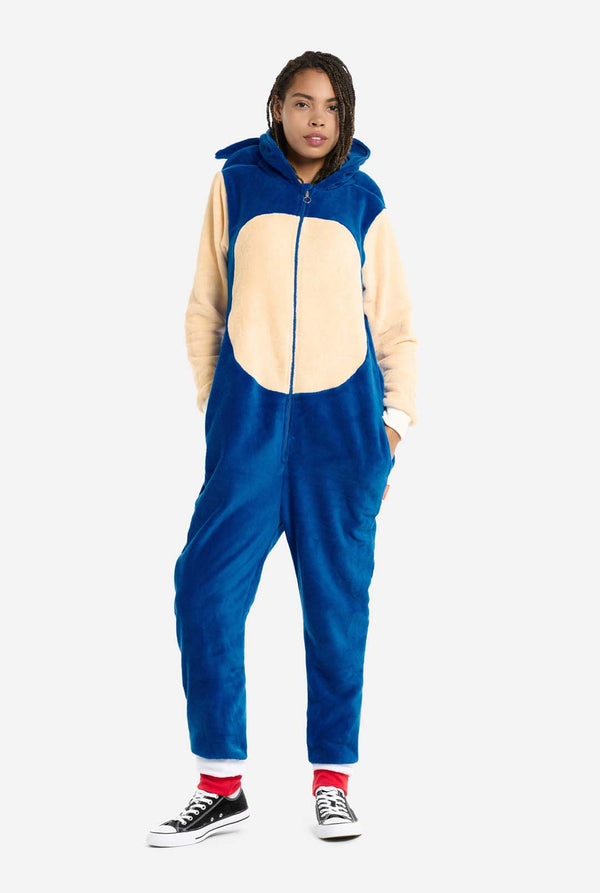 Eine Frau trägt einen kuscheligen, blauen Sonic the Hedgehog-Onesie für Erwachsene. Weiche beigefarbene Patches auf der Vorderseite und an den Ärmeln imitieren Sonics ikonischen Look. Die Kapuze ist mit Sonics Gesicht gestaltet. Der Reißverschluss vorne sorgt für einen bequemen Sitz. Die Frau steht mit den Händen in den Seitentaschen des Onesies. Ihr Gesichtsausdruck ist ruhig und entspannt. Dieser Sonic the Hedgehog-Onesie ist ideal für Cosplay, zum Entspannen oder für Themenveranstaltungen.