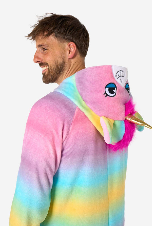 Ein Mann trägt einen farbenfrohen Einteiler für Erwachsene in Form eines Regenbogen-Einhorns. Der Einteiler besticht durch einen leuchtenden Farbverlauf in Pastellfarben, ein goldenes Horn und verspielte Einhorn-Details – perfekt für gemütliches Faulenzen oder Kostümpartys. Die Kapuze liegt flach auf seinem Rücken. Nahaufnahme des Oberkörpers (Seitenansicht).
